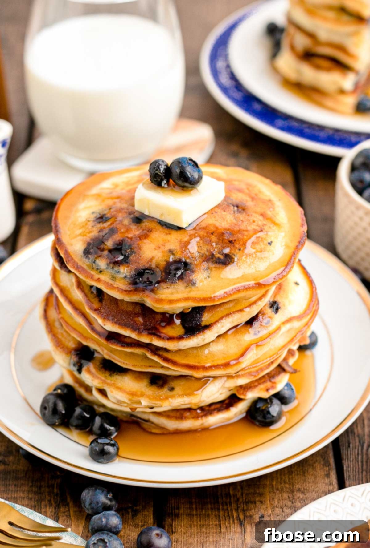 A stack of blueberry pancakes on a white plate on a wooden table with a glass of milk in the background. 