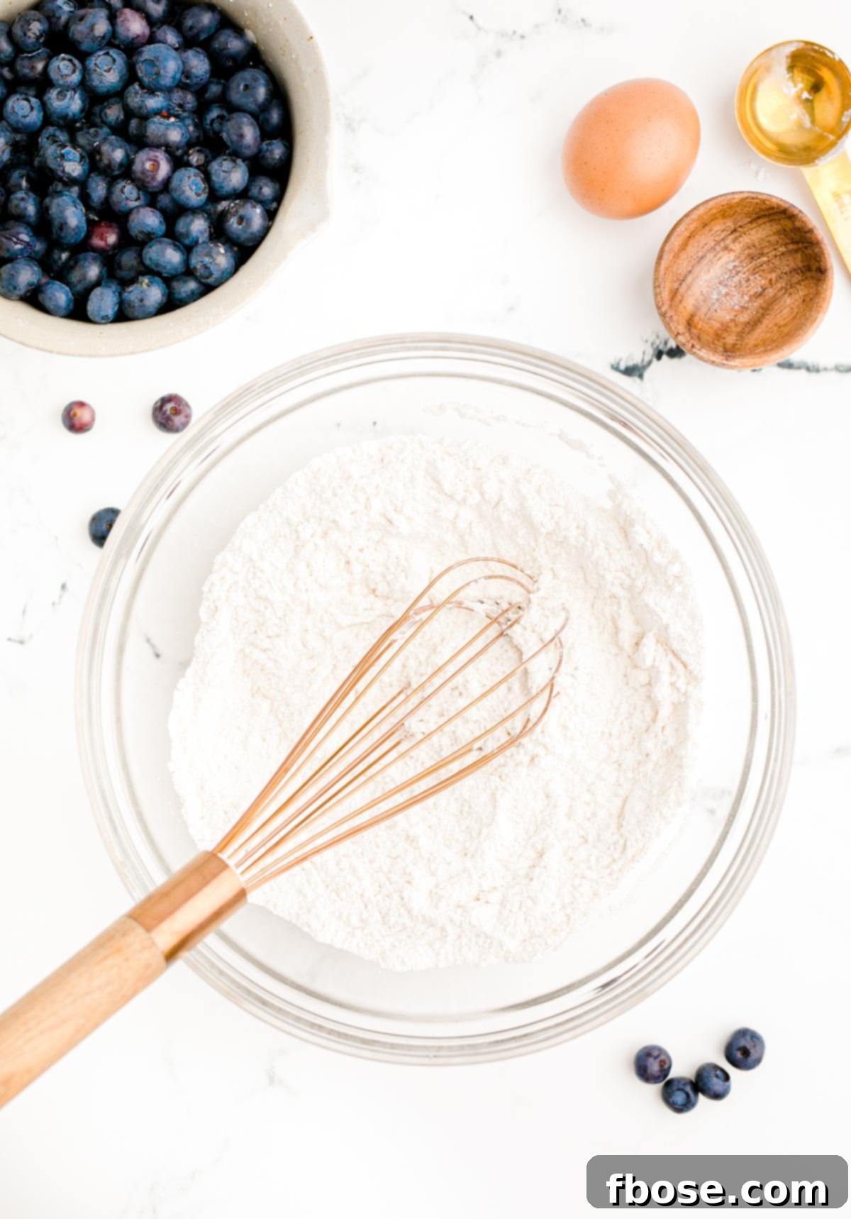 Overhead photo of dry ingredients being mixed in a glass bowl to make blueberry pancakes on a white table.