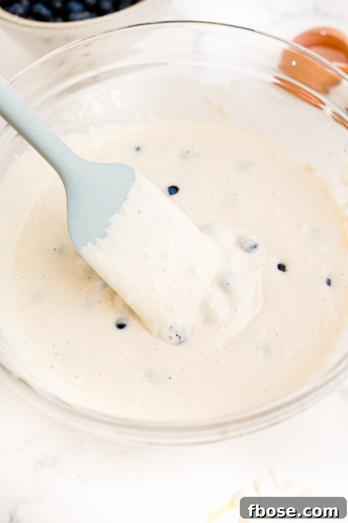 Blueberry pancake batter in a glass mixing bowl with a blue rubber spatula.