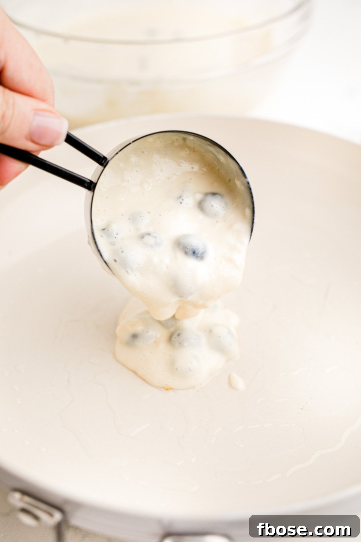 A measuring cup pouring blueberry pancake batter onto a white skillet.
