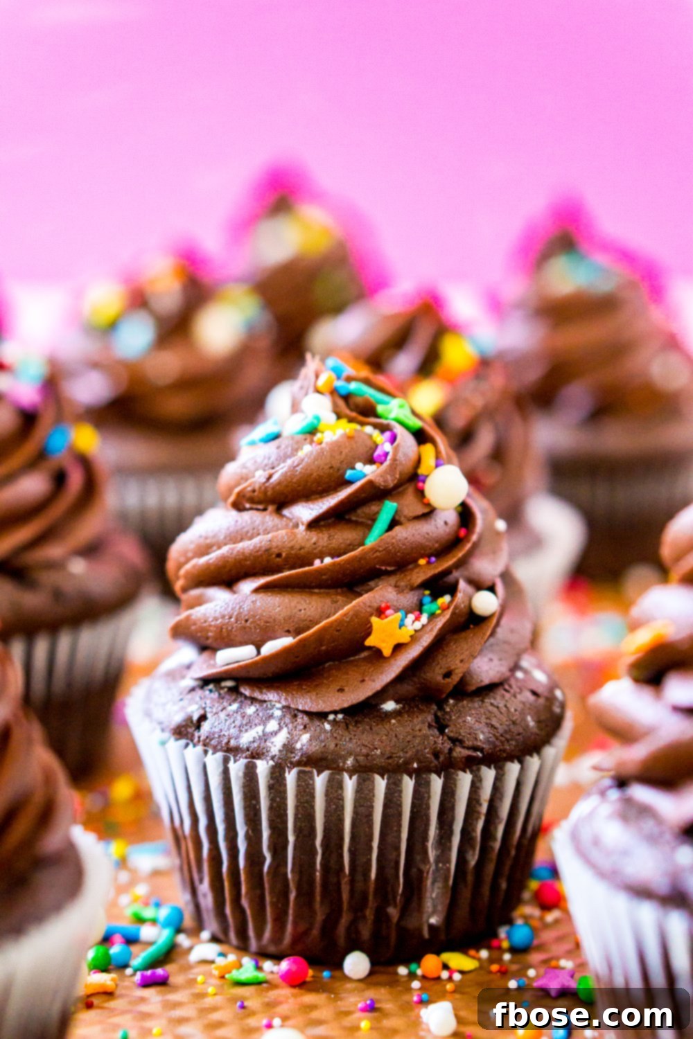 Super moist Chocolate Cupcakes cooling on a baking sheet, adorned with vibrant rainbow colored sprinkles and set against a soft pink background.
