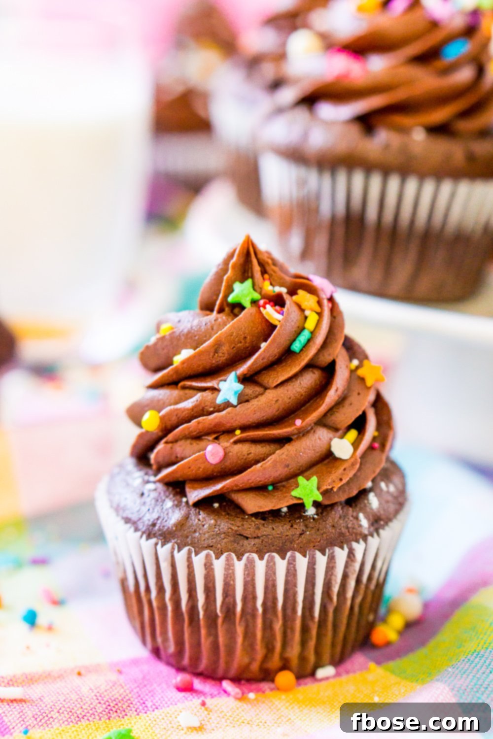 A delectable Chocolate Cupcake presented on a colorful napkin, with a pristine white cake stand holding an array of other chocolate cupcakes in the background, ready for serving.