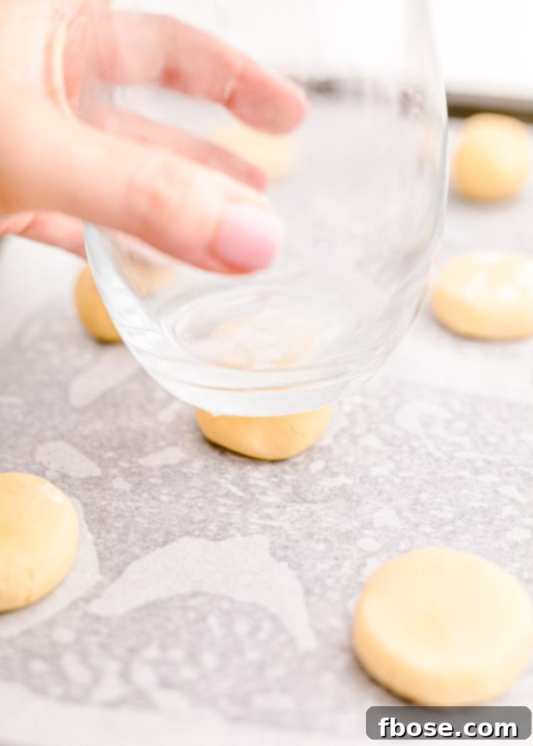 Festive Fir Tree Treats 8 A woman's hand pressing down on balls of sugar cookie dough with the flat bottom of a glass, transforming them into uniform, flat discs on a baking sheet.