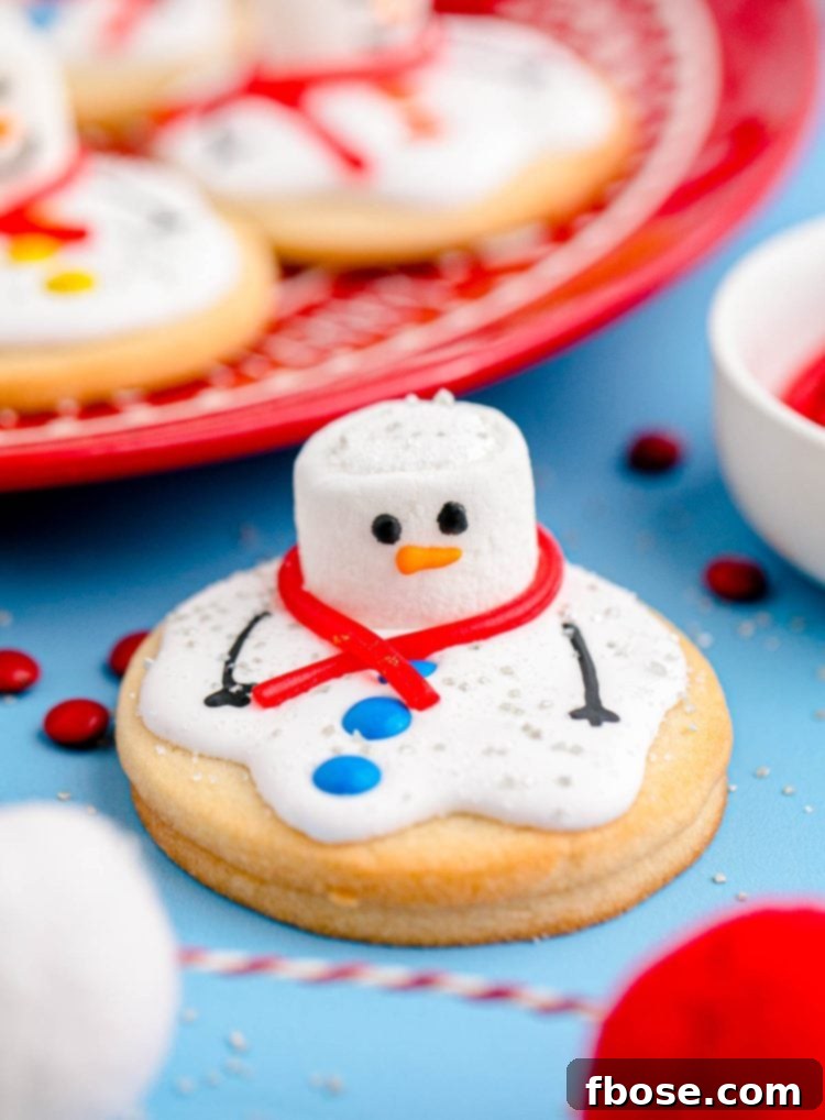 Melty Snowman Cookies 2 Close up photo of melted snowman cookies on a blue counter with some on a red plate in the background. Each cookie features a marshmallow head, candy buttons, and royal icing 'puddle'.