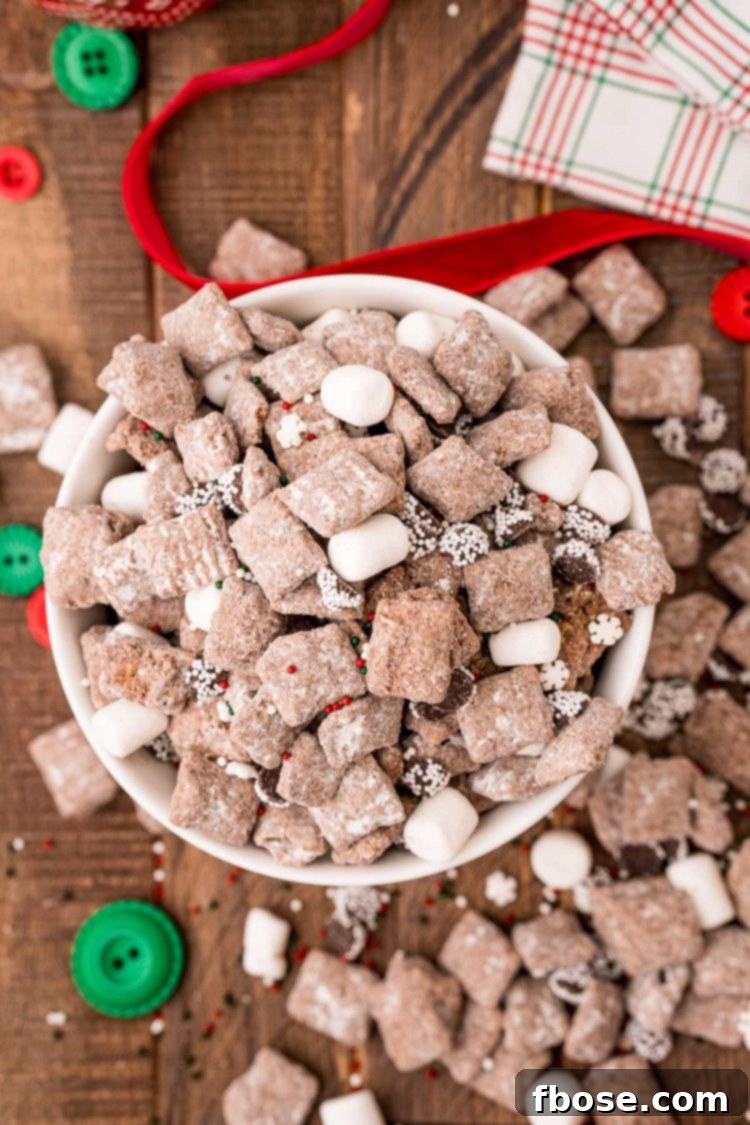 Overhead photo of a bowl of hot chocolate muddy buddies on a wooden table with scattered holiday decorations like pinecones and cinnamon sticks.