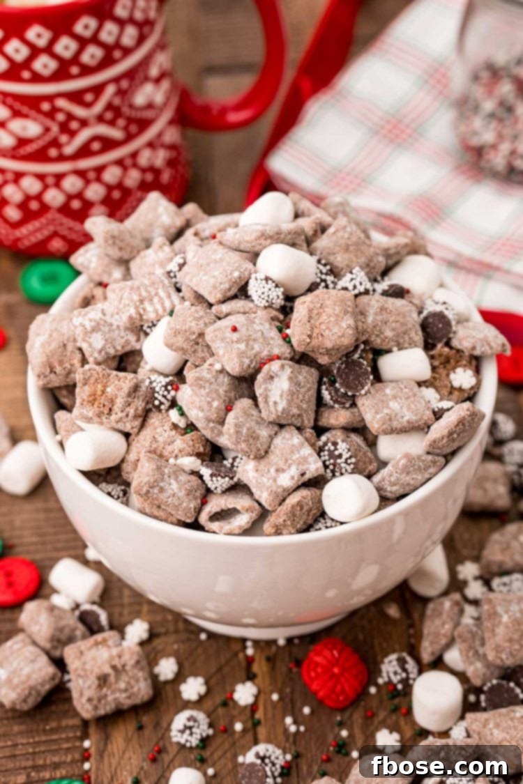 A white bowl filled with hot chocolate muddy buddies on a wooden table, with loose muddy buddies and holiday decorations like pinecones and small gifts scattered around.