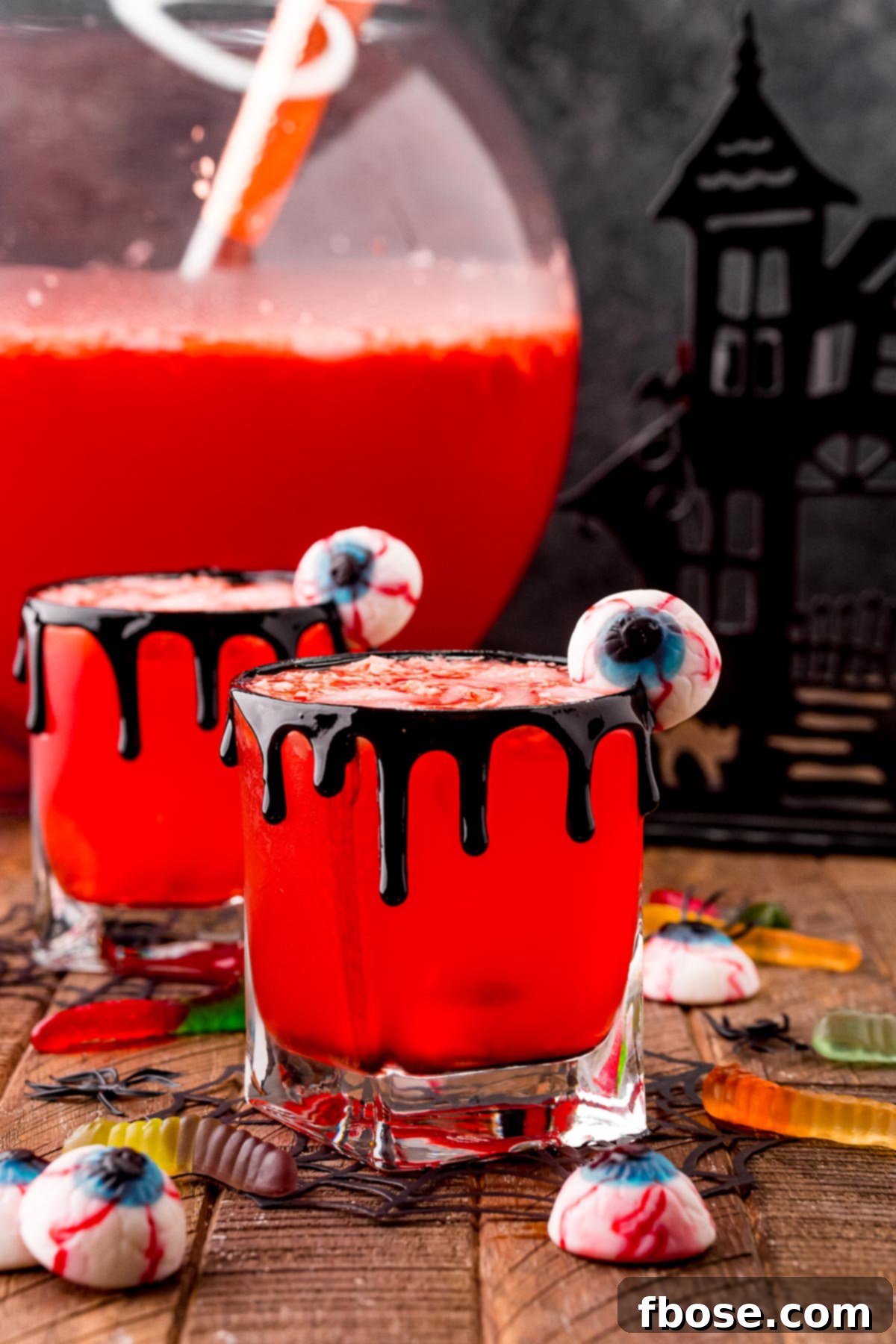 Two glasses filled with vibrant red Werewolf Halloween punch, garnished with gummy worms and candy eyeballs, resting on a rustic wooden table. In the background, a large punch bowl is brimming with the festive drink, surrounded by additional gummy candies, setting a perfectly spooky scene.