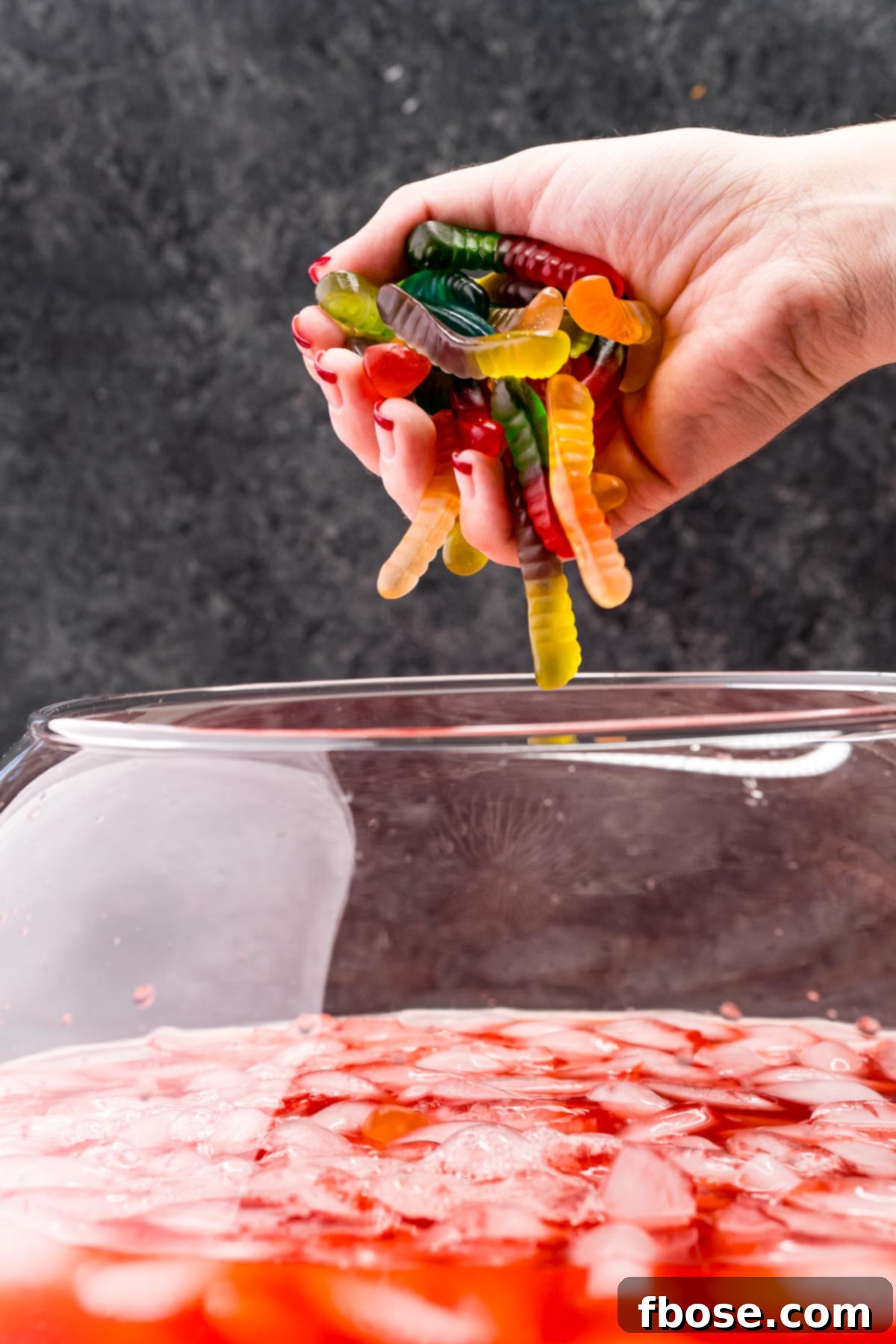 A woman's hand gracefully drops a handful of vibrant gummy worms into a punch bowl filled with sparkling red Halloween punch, ready to add a spooky, playful touch to the beverage.