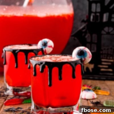 Photo of two glasses with Werewolf Halloween punch in them on a wooden table surrounded but gummy worms and eyeball gummies with a large punch bowl in the background.