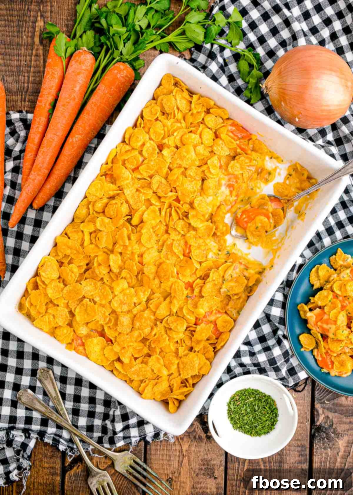 Overhead photo of a white casserole dish filled with carrot casserole on a black and white checkered napkin. The golden cornflake topping is clearly visible.
