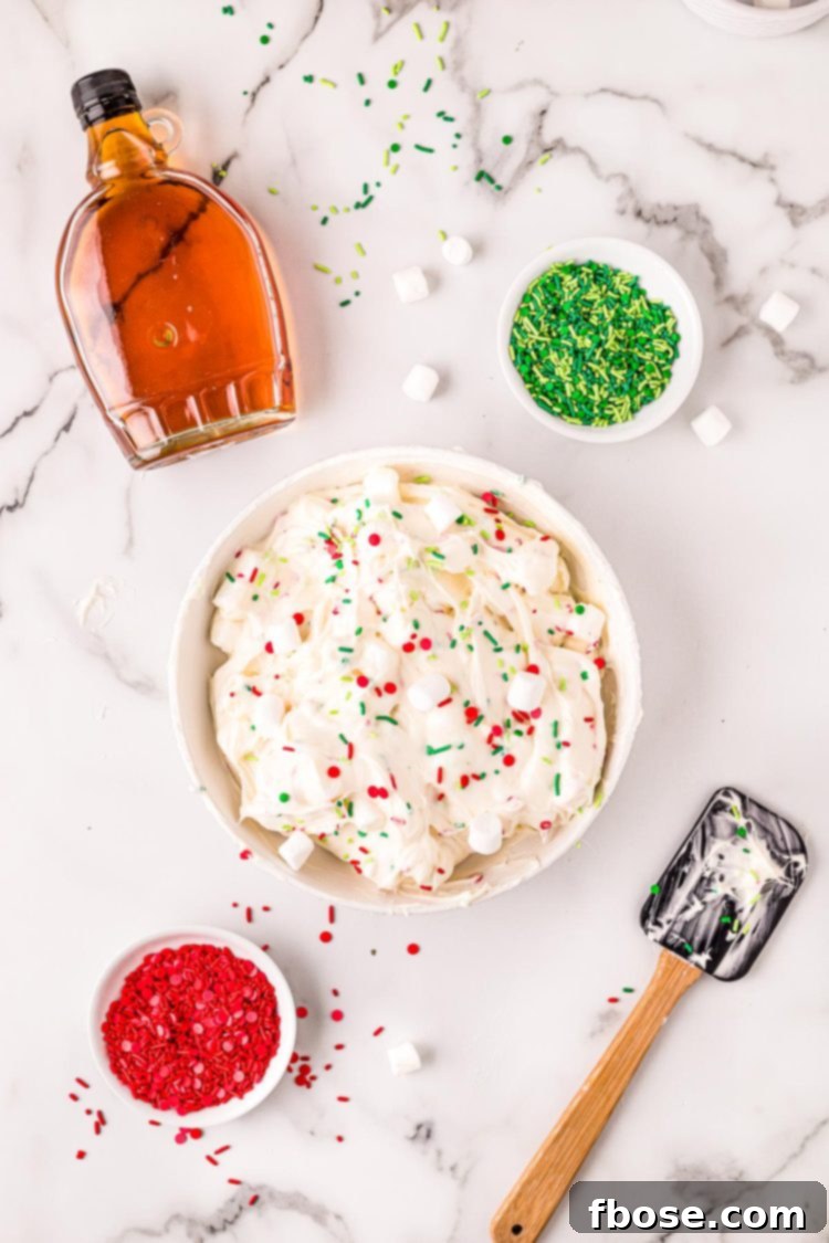 Overhead photo of Christmas dessert dip in a white bowl on a marble surface with a rubber spatula, maple syrup, and bowls of sprinkles around it.
