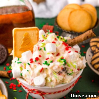 Close up photo of a white bowl filled with Christmas dessert dip with cookies around it on a green surface.