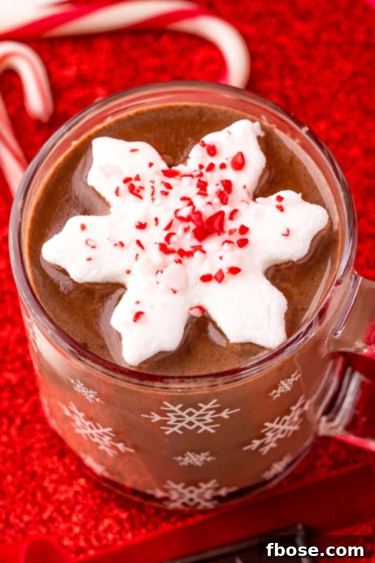 Close up photo of a mug of peppermint hot chocolate on a red surface with candy canes and chocolate and red ribbon around it.