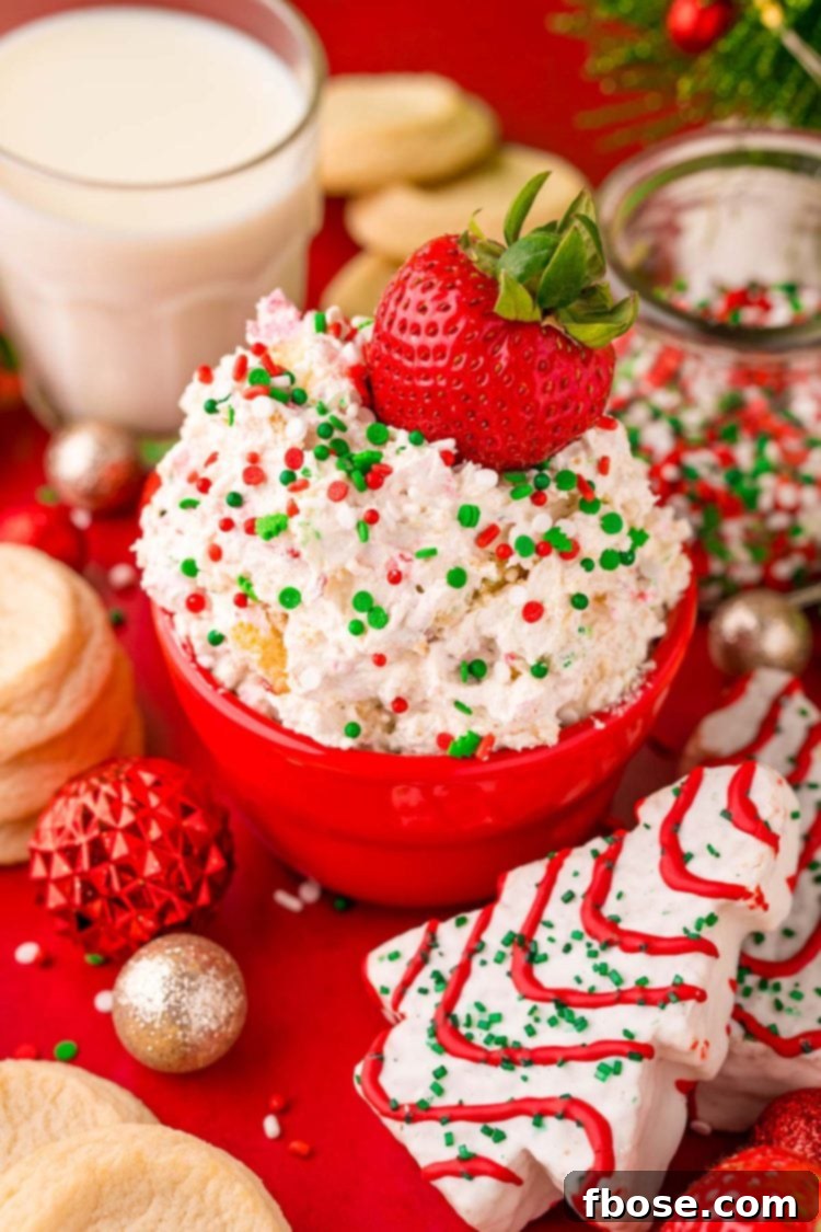 Close up photo of Little Debbie Christmas Tree Cake Dip in a festive red bowl on a red surface, surrounded by holiday decorations, with a fresh strawberry dipped into it.