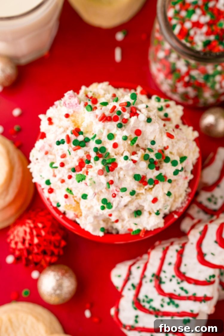 Close up photo of Christmas Tree Cake Dip in a red bowl on a red surface with holiday decorations scattered around, emphasizing its festive appeal.