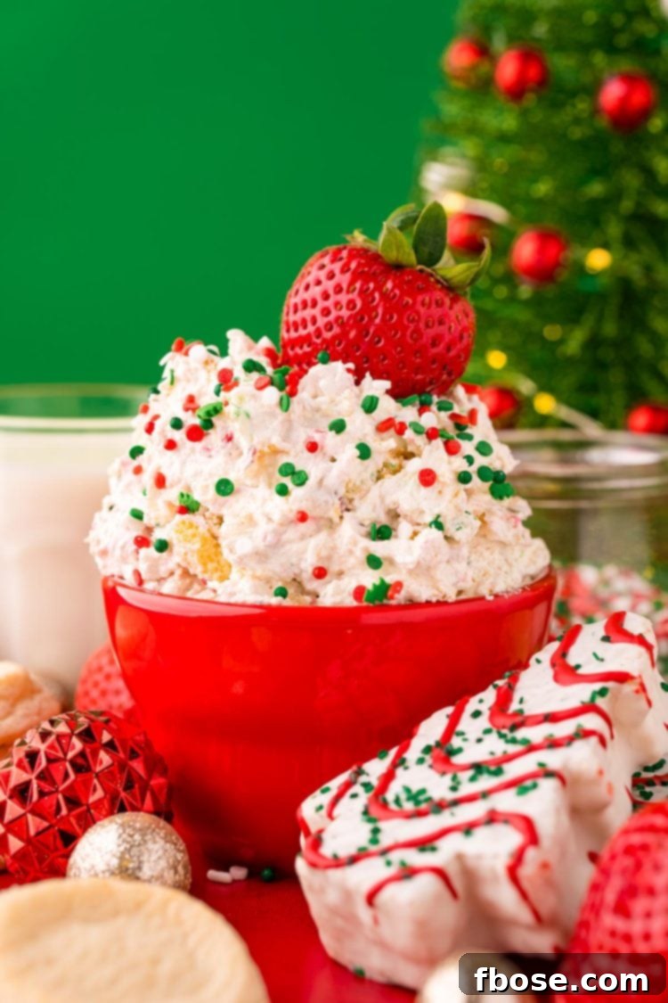 Close up photo of Little Debbie Christmas Tree Cake Dip in a red bowl on a red surface surrounded by holiday decorations, with a fresh strawberry dipped into it, highlighting its irresistible appeal.