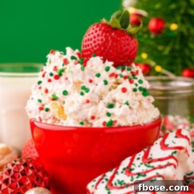 Close up photo of Little Debbie Christmas Tree Cake Dip in a red bowl on a red surface surrounded by holiday decorations with a strawberry dipped into it.