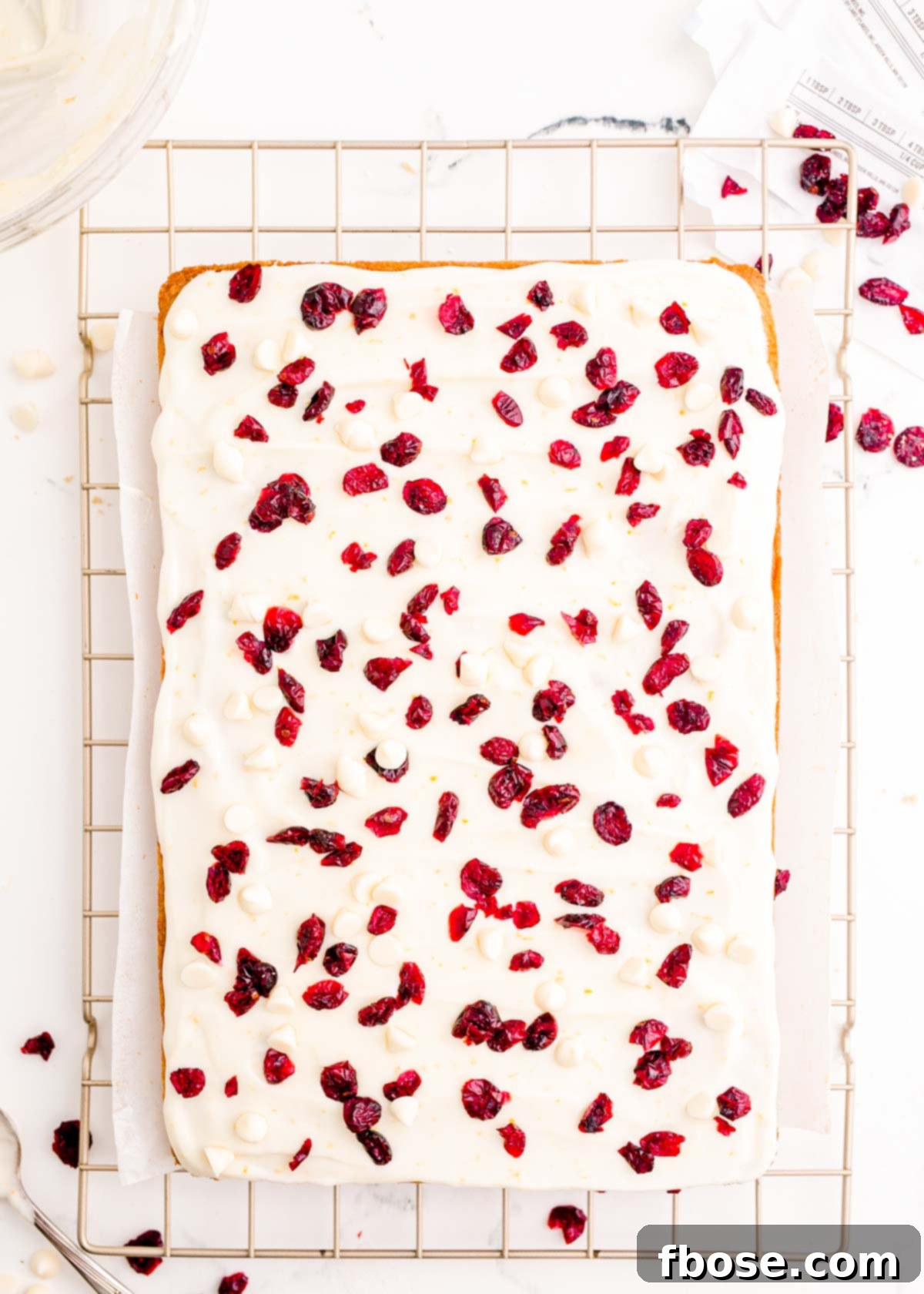 Deliciously frosted cranberry bliss bars, now garnished with a sprinkle of cranberries and white chocolate chips, await their final drizzle on a wire rack.