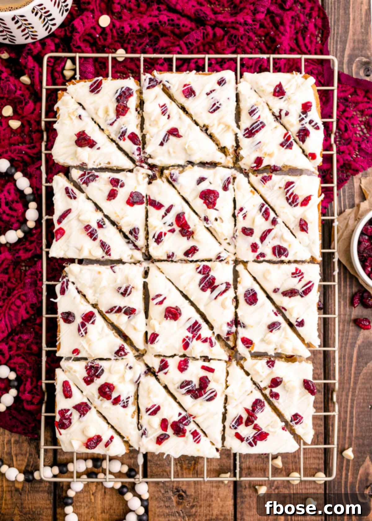 Overhead photo of fresh homemade cranberry bliss bars cooling on a wire rack set on a rustic wooden table with a festive red lace napkin nearby.