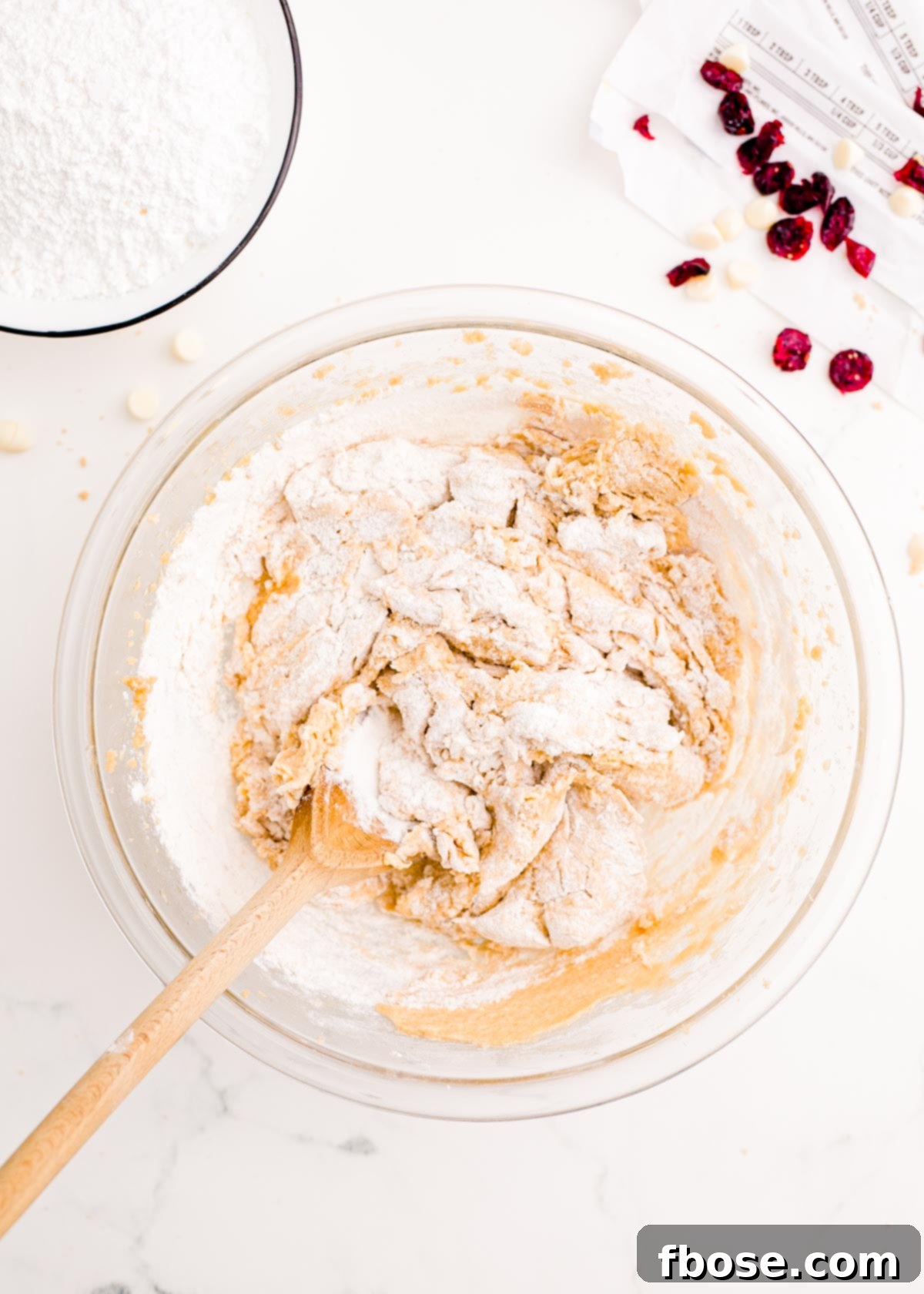 The dry ingredients are being gently mixed into the wet ingredients in a large glass mixing bowl, forming the base for the cranberry bliss bars.