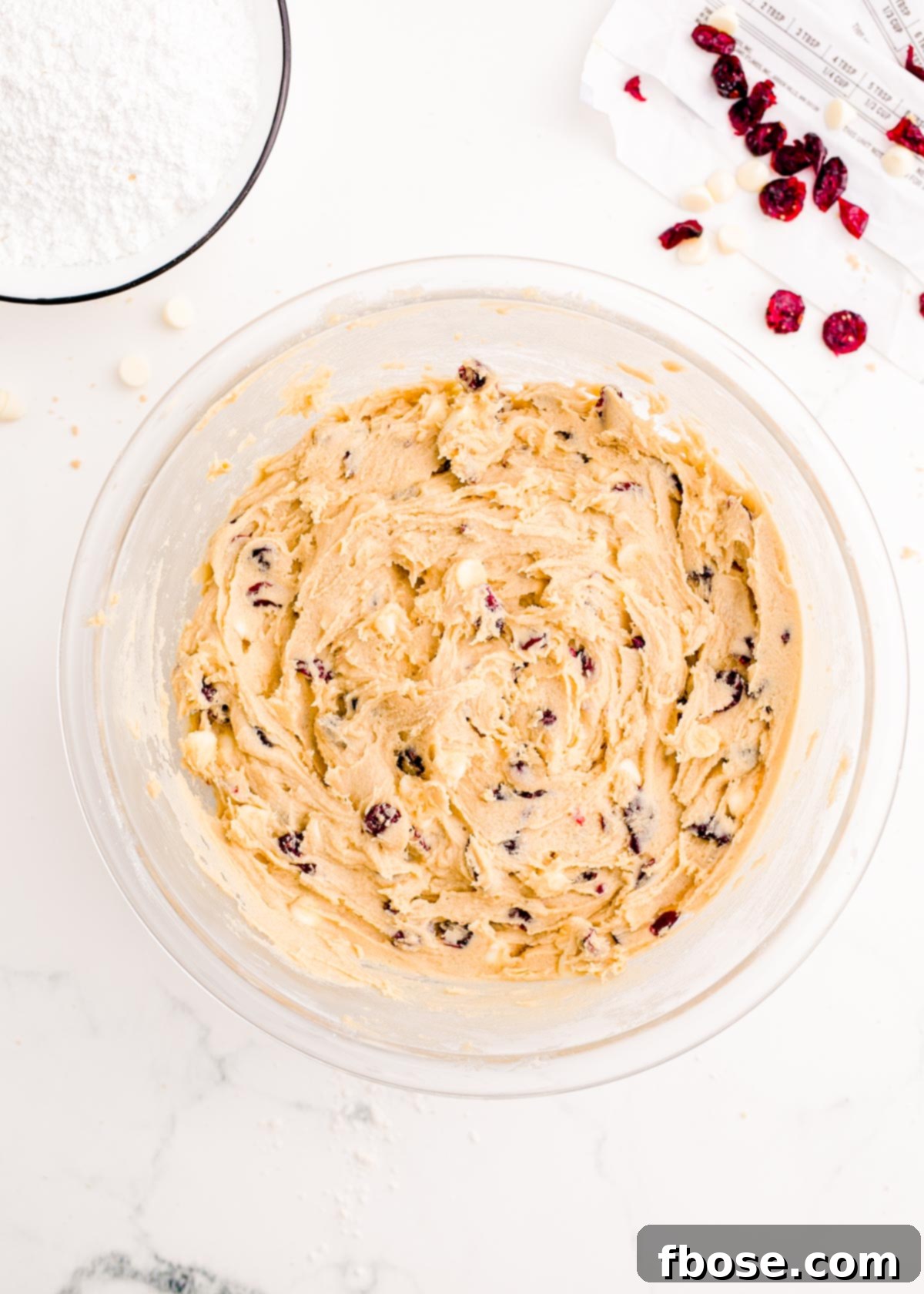 The complete batter for cranberry bliss bars, studded with white chocolate chips and dried cranberries, rests in a large glass mixing bowl.