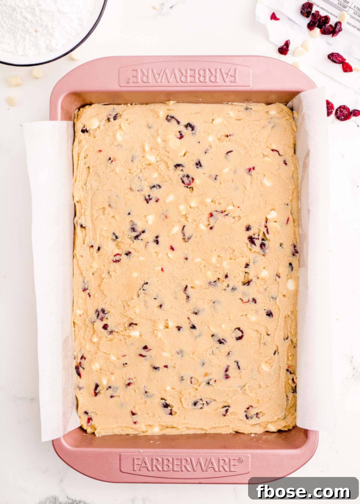 Cranberry bliss bar dough, vibrant with red cranberries and white chocolate, neatly spread in a pink baking pan lined with parchment paper, ready for baking.