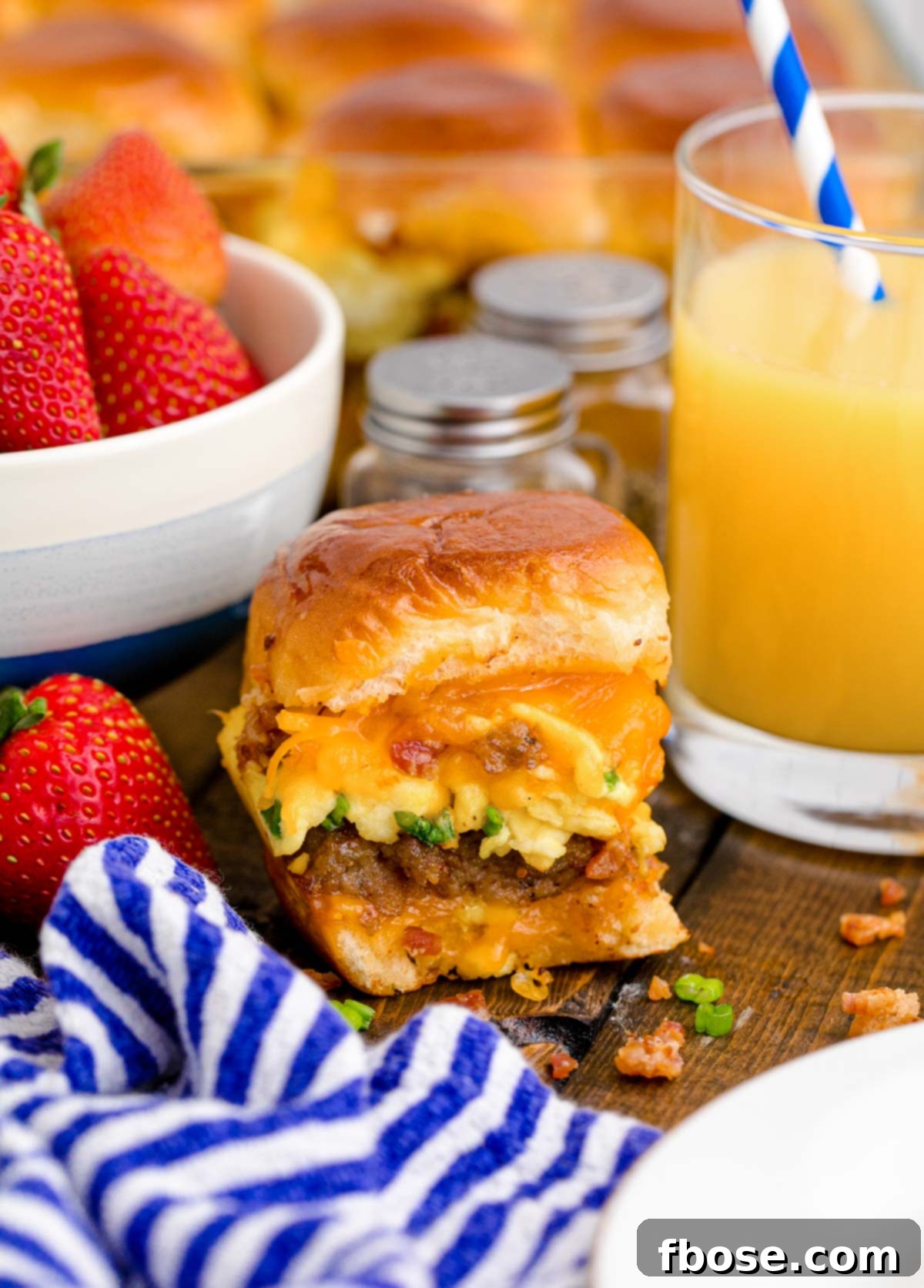 Close up photo of breakfast sliders on a wooden table with a glass of orange juice and bowl of strawberries around it.