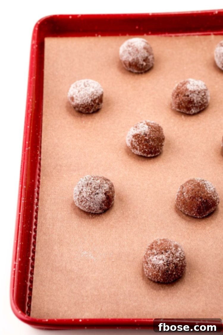 Mint Chocolate Thumbprint Treasures 6 Balls of chocolate cookie dough rolled in granulated sugar on a red parchment-lined baking sheet.