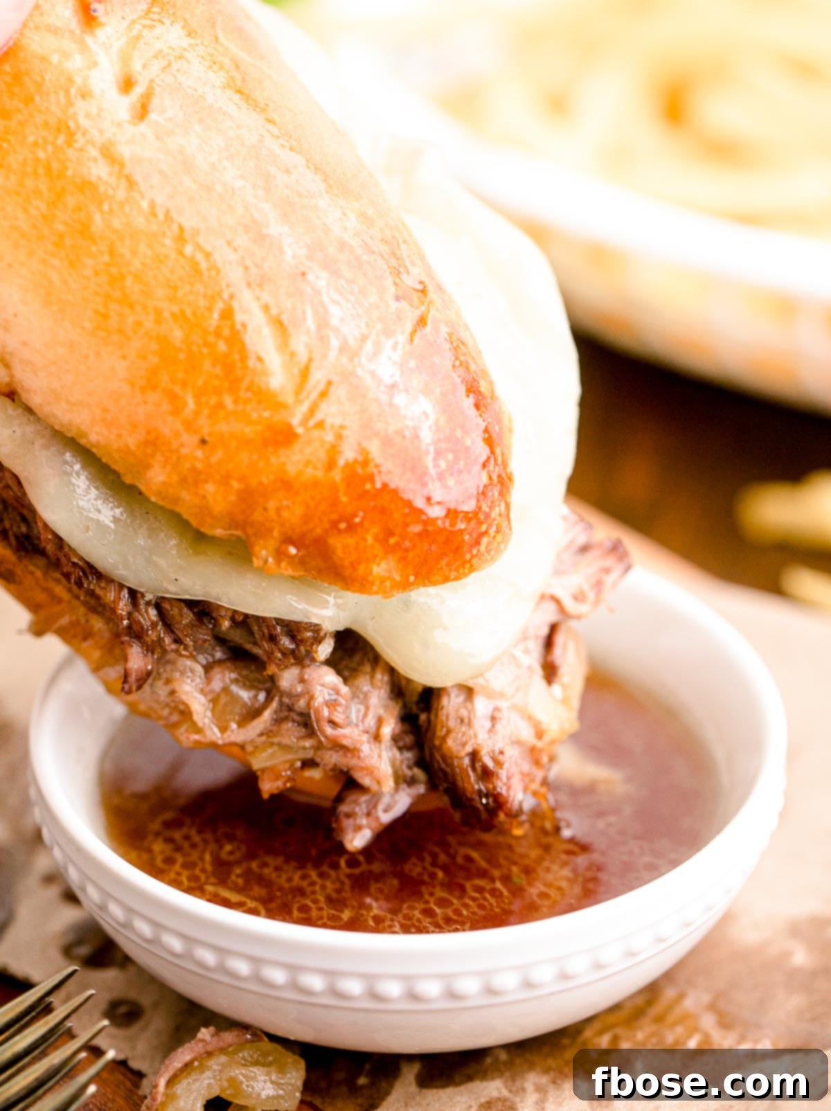 A French Dip Sandwich being dipped into a small bowl of savory au jus, highlighting the rich dipping experience.
