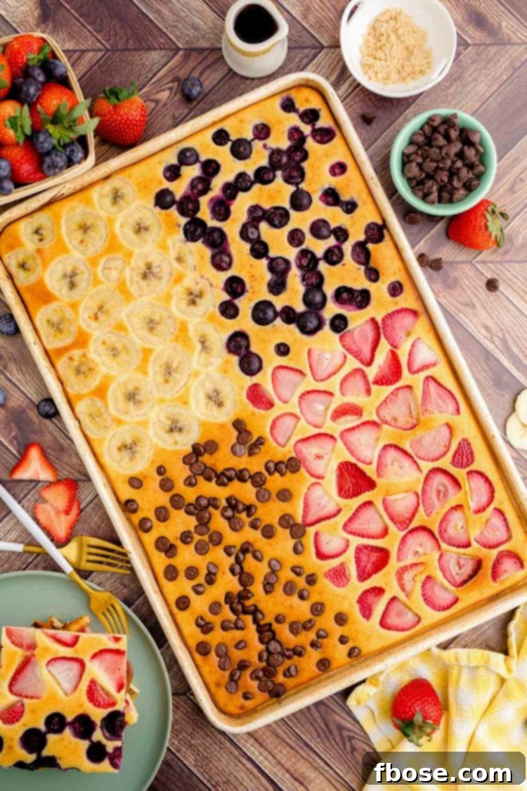 Overhead view of a baked sheet pan pancake divided into four sections, each with different toppings like blueberries, chocolate chips, strawberries, and bananas, ready to be served.