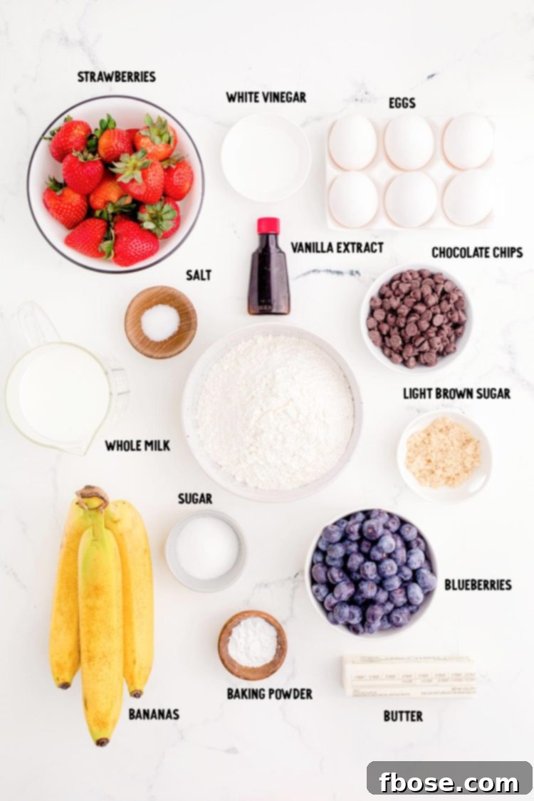 Assortment of fresh ingredients for sheet pan pancakes laid out on a marble surface, including flour, sugar, eggs, milk, vanilla, butter, blueberries, and chocolate chips.