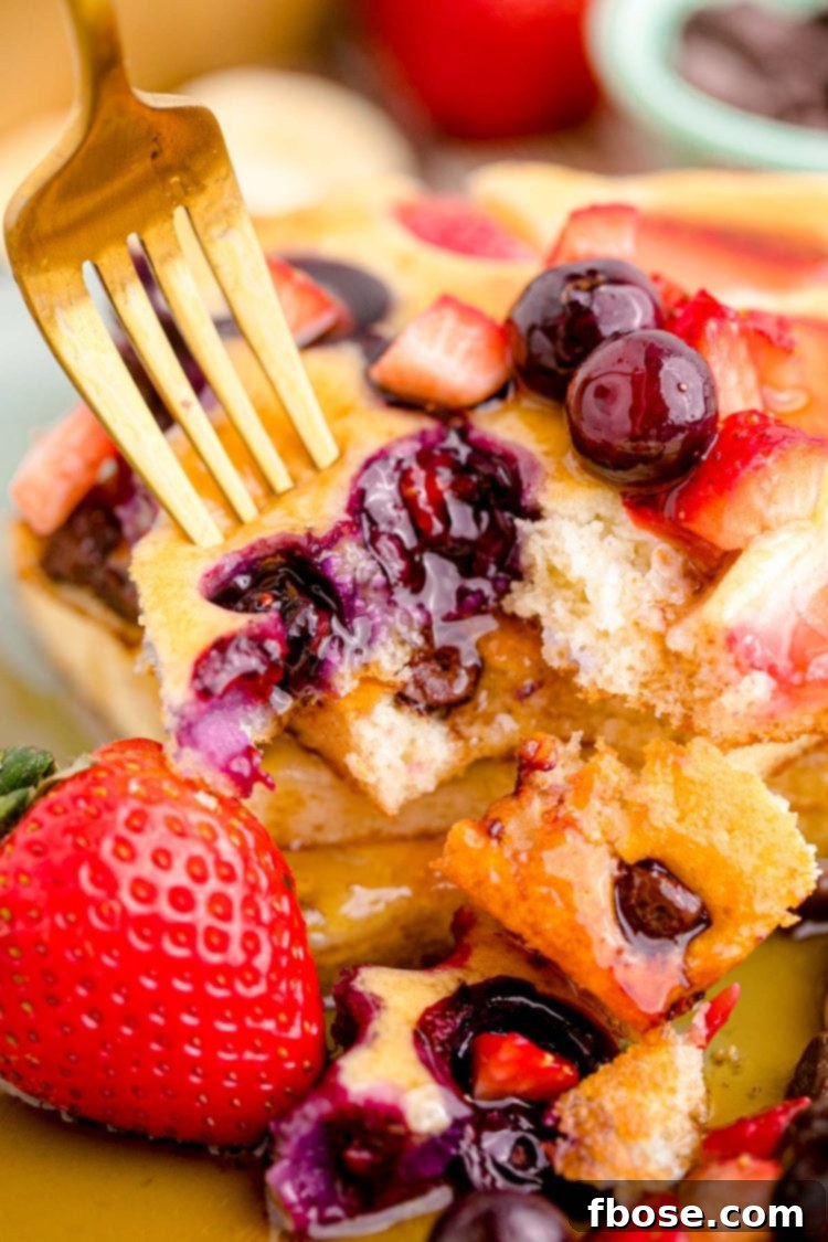 A fork lifts a bite of sheet pan pancake from a stack, revealing its fluffy interior and delicious blueberry and strawberry mix-ins.