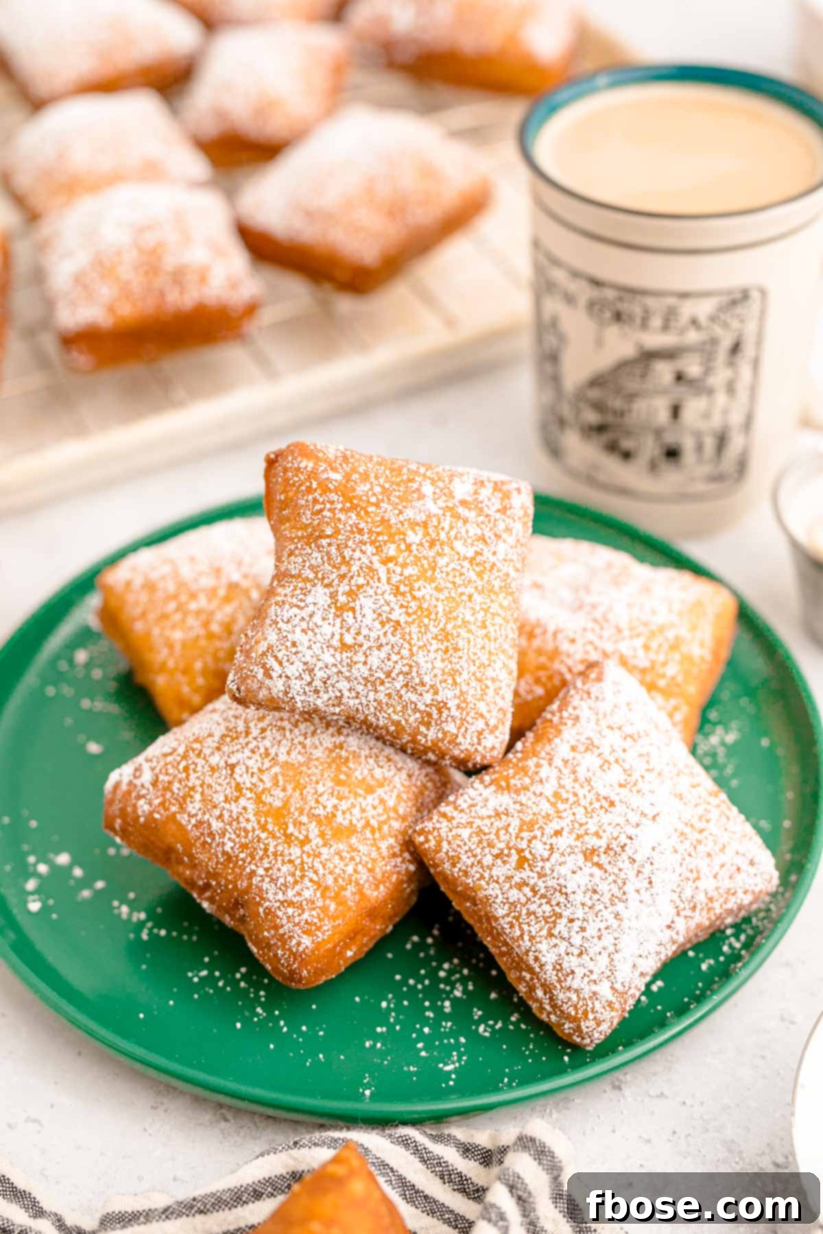 Close up photo of beignets on a green plate with a mug of coffee and more beignets in the background.