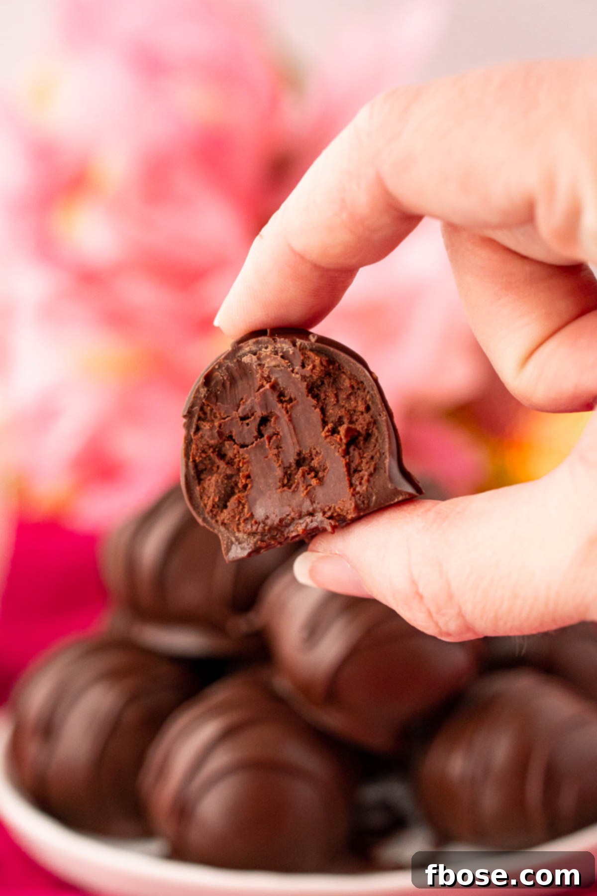 The Ultimate Chocolate Truffles Recipe 6 Close up photo of a woman's hand holding a chocolate truffle with a bite taken out of it. More truffles on a plate in the background.