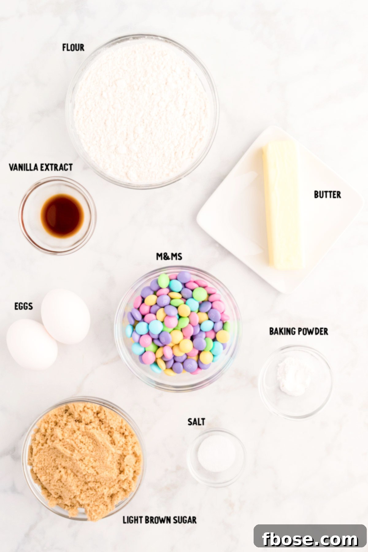 A flat lay photograph showcasing all the fresh ingredients neatly arranged on a marble counter, ready for making Easter blondies, including butter, sugar, eggs, flour, and pastel M&Ms.