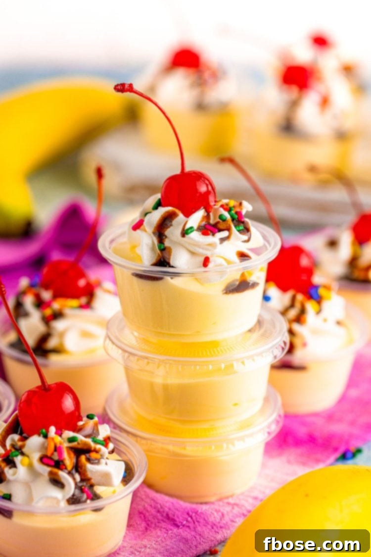 A stack of three banana split pudding shots topped with whipped cream, chocolate syrup, and a maraschino cherry on a white surface.