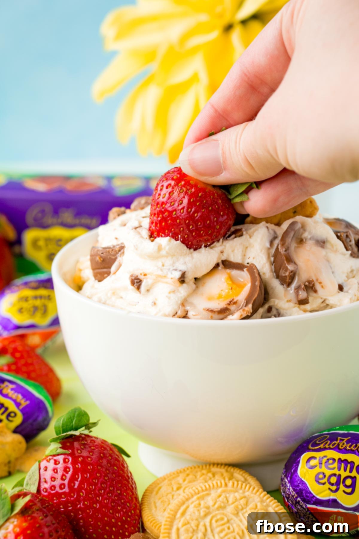 A woman's hand dipping a fresh strawberry into a bowl of creamy Cadbury Creme Egg Dip.