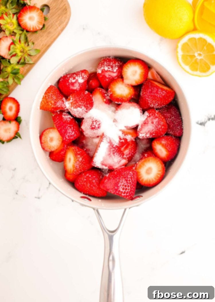 Fresh strawberries, a bowl of granulated sugar, and a small glass of lemon juice arranged on a white marble counter.