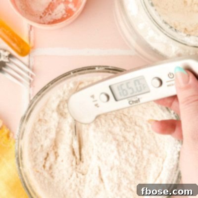 flour in a glass bowl with a woman's hand taking it's temperature with a kitchen thermometer.