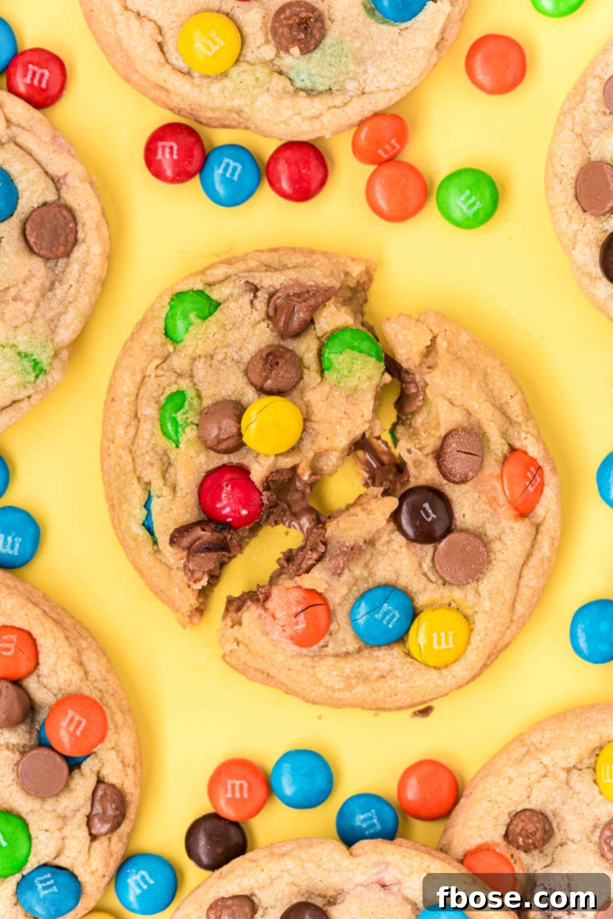 A vibrant close-up of M&M cookies on a yellow surface, with one cookie playfully broken in half to reveal its soft, chewy interior and abundant mix-ins.