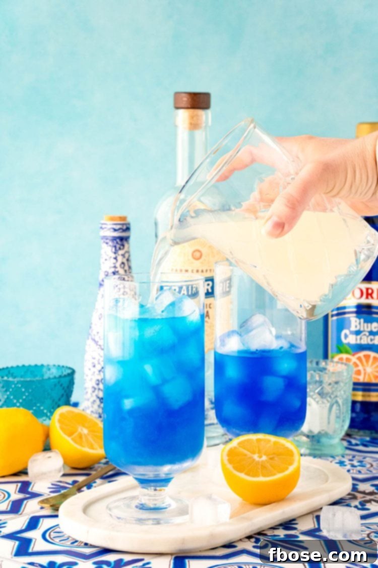 Azure Escape 5 Fresh lemonade cascades from a clear glass pitcher into a tall glass filled with ice, setting the stage for mixing a beautiful blue cocktail.
