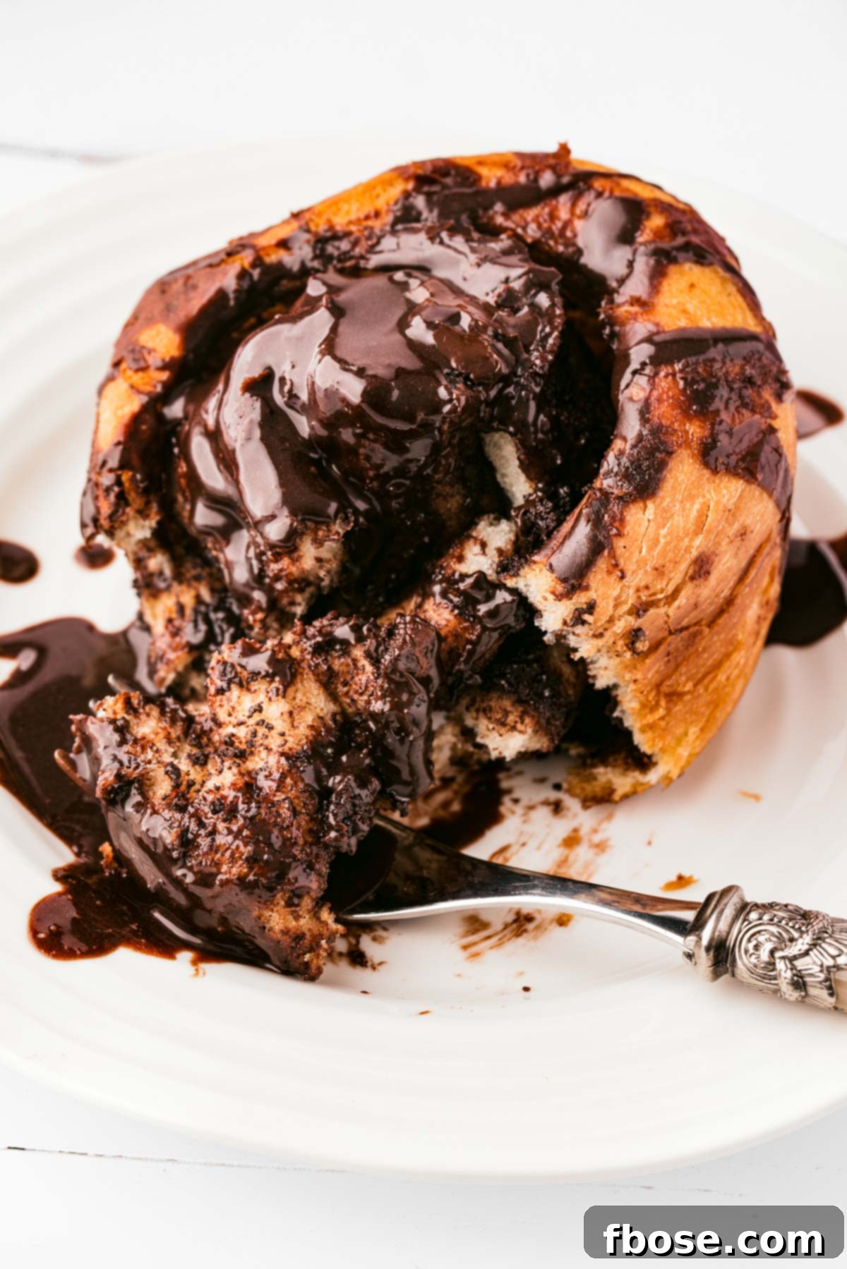 Decadent Chocolate Cinnamon Swirls 18 A chocolate cinnamon roll on a white plate with a fork taking a bite out of it.