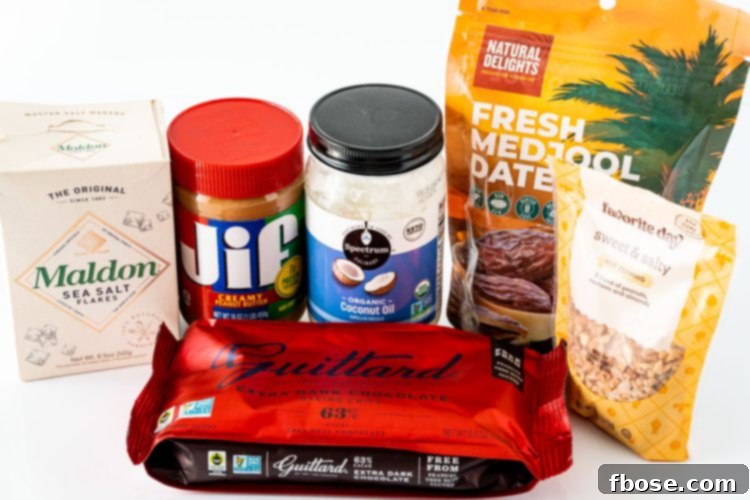 A collection of ingredients laid out on a white surface, ready to make Snickers Stuffed Dates: Medjool dates, peanut butter, chopped peanuts, dark chocolate chips, and coconut oil.