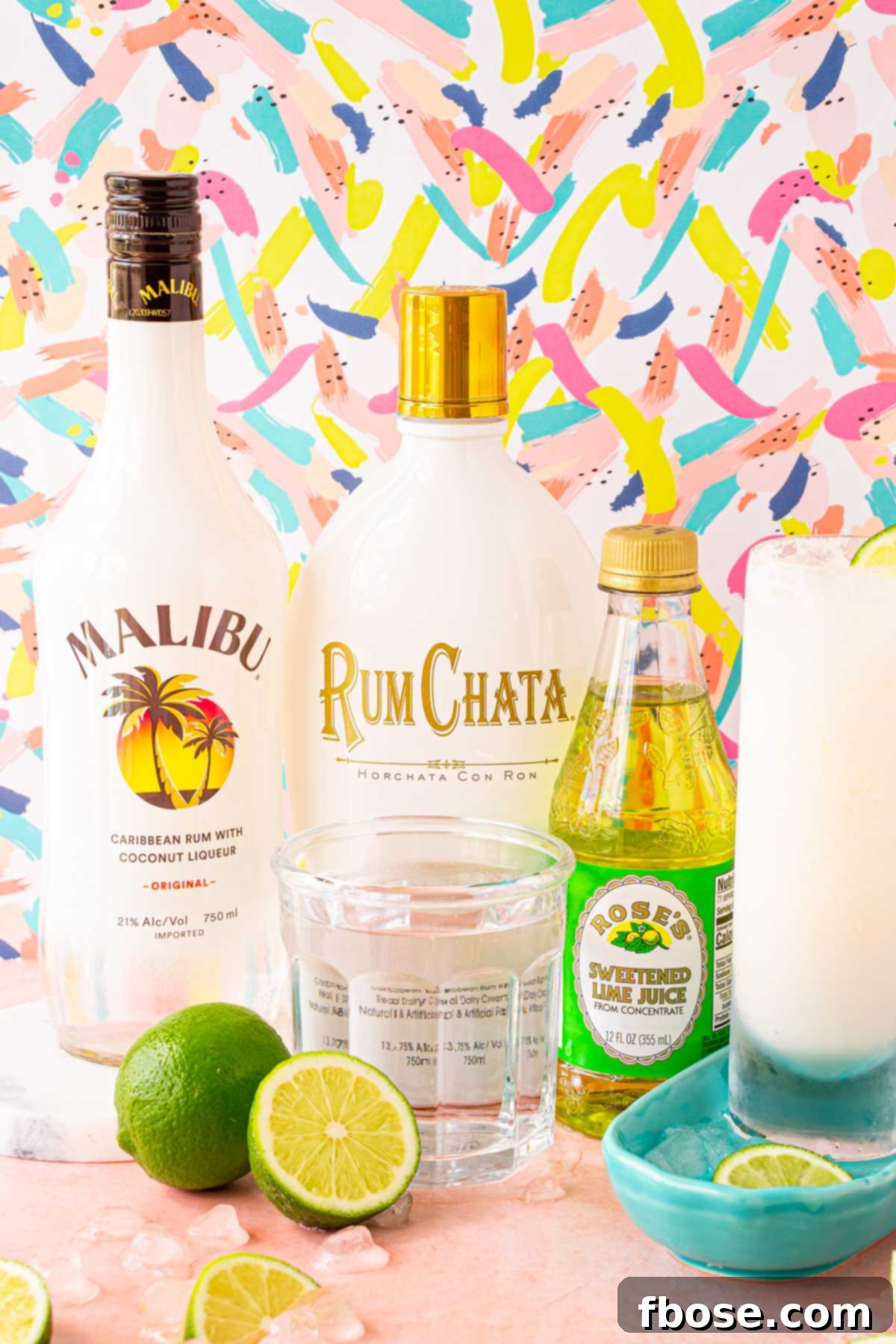 Malibu, RumChata, Lime Juice bottles and a glass of club soda on a pink surface surrounded by fresh limes.