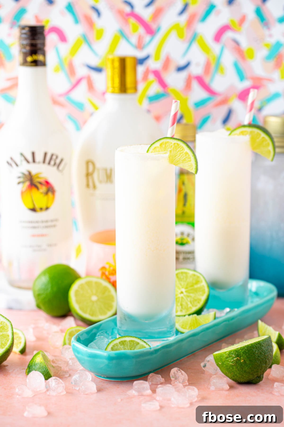 Two glasses with white cocktails on a teal dish, surrounded by limes and ice, ready to be served.
