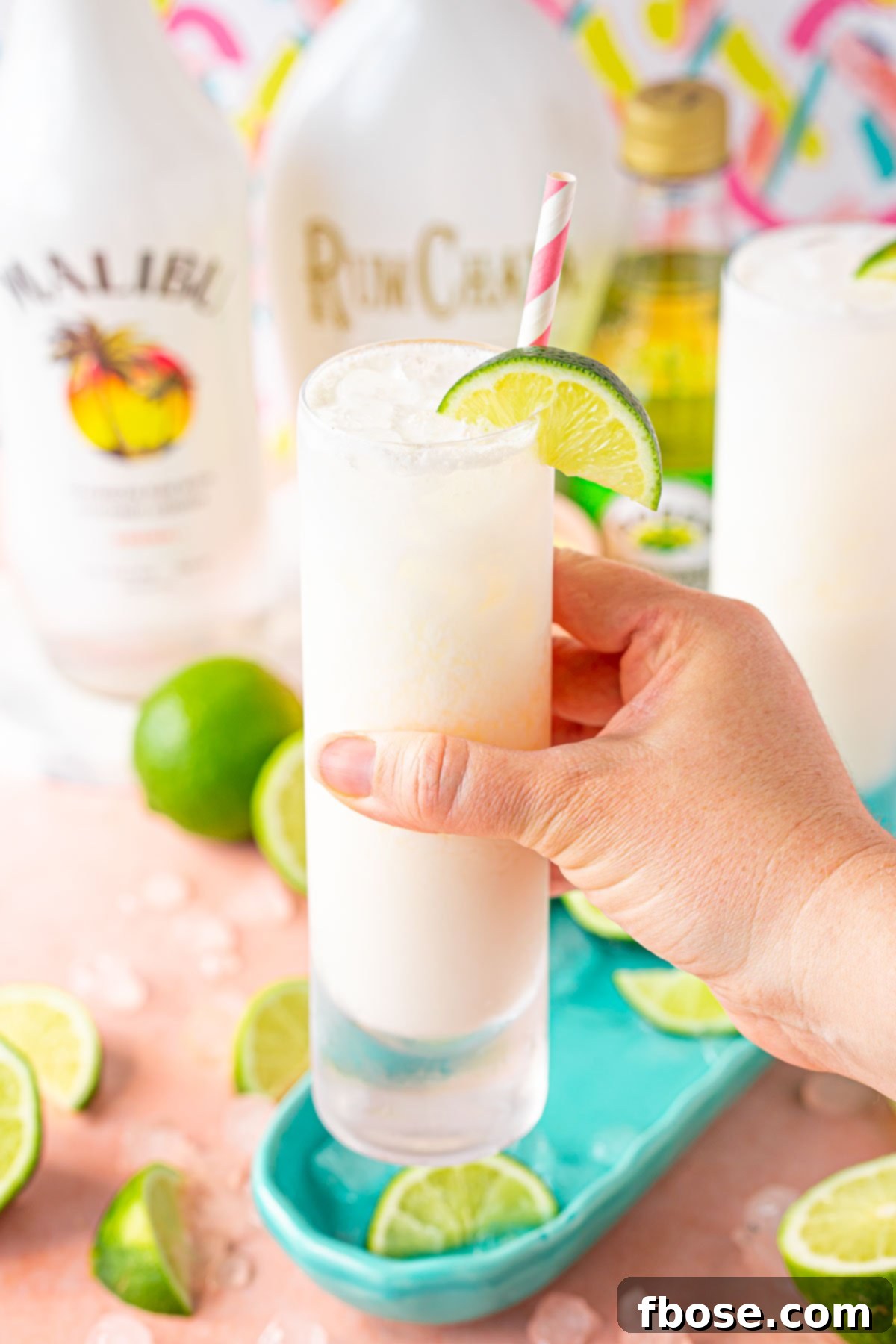 A woman's hand holding up a RumChata Malibu Lime and Soda cocktail in a tall glass, with a lime wedge garnish, to the camera.