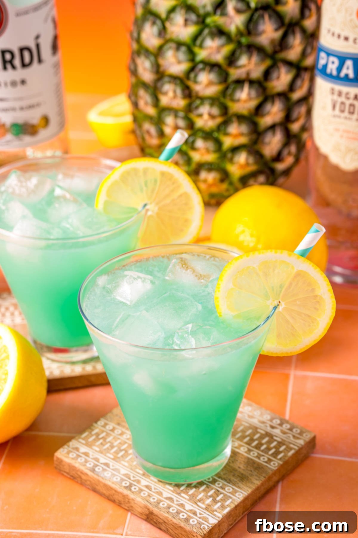 Mermaid's Paradise Elixir 6 Two glasses of vibrant blue Mermaid Rum Punch, garnished with a lime wedge, resting on a bright orange outdoor table, hinting at a fun summer day.