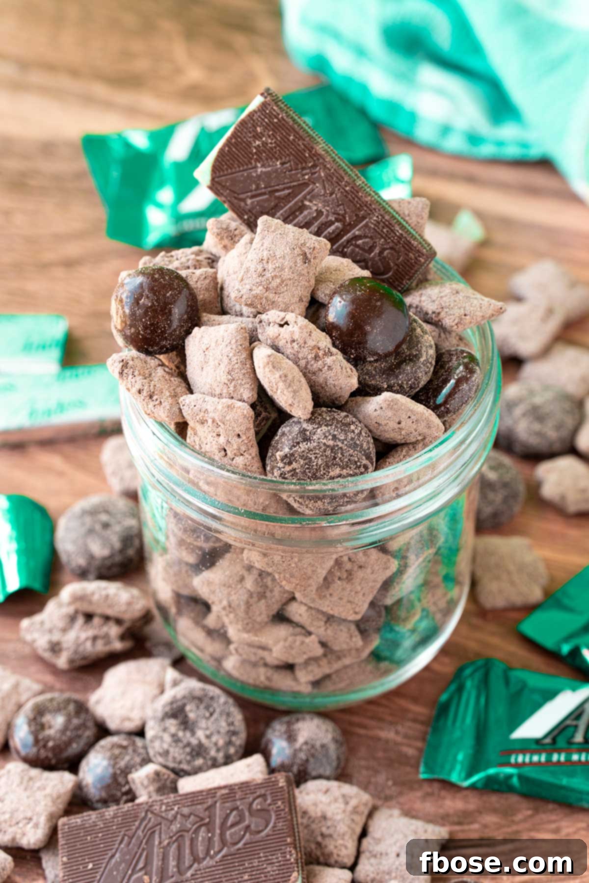 No-Bake Mint Chocolate Muddy Buddies 2 Mint chocolate muddy buddies in a jar on a wooden table surrounded my muddy buddies and mint candies.