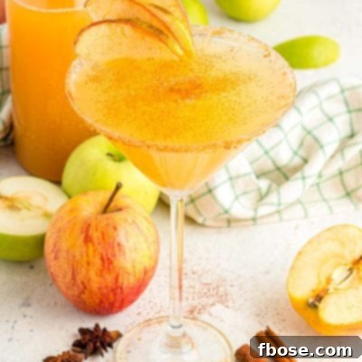 Apple cider martini surrounded by apples, cinnamon sticks, and a bottle of cider.