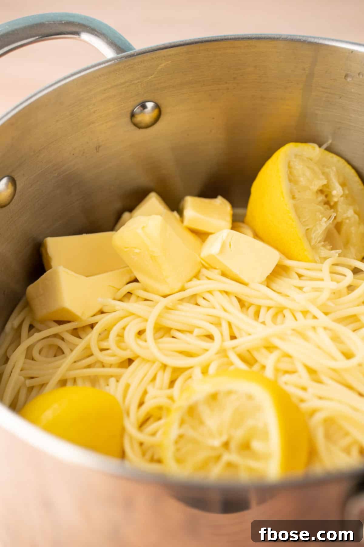 Spaghetti with lemon halves, lemon juice, and butter cubes in a stainless steel pot, beginning to emulsify.